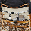 A beige canvas crossbody bag with a leather strap and hanging ring is shown on a wooden folding chair.