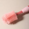 A pink silicone baby bottle cleaning brush with bristles is shown against a light background.