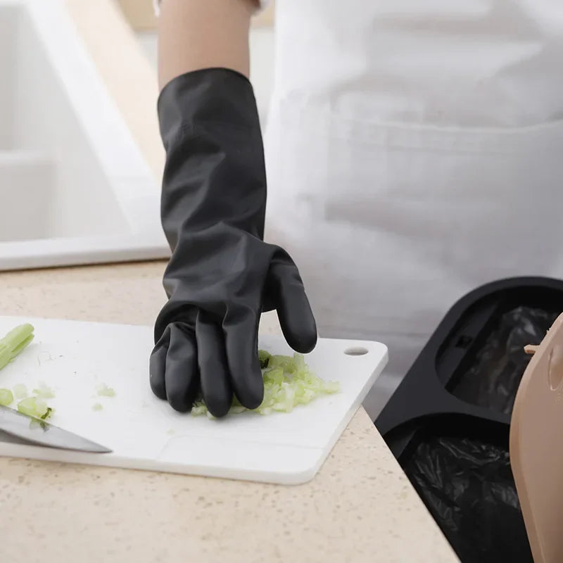 A hand wearing a black durable rubber cleaning glove is holding chopped green vegetables on a white cutting board.