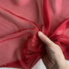A hand is folding a piece of sheer, reddish-pink silk chiffon fabric.