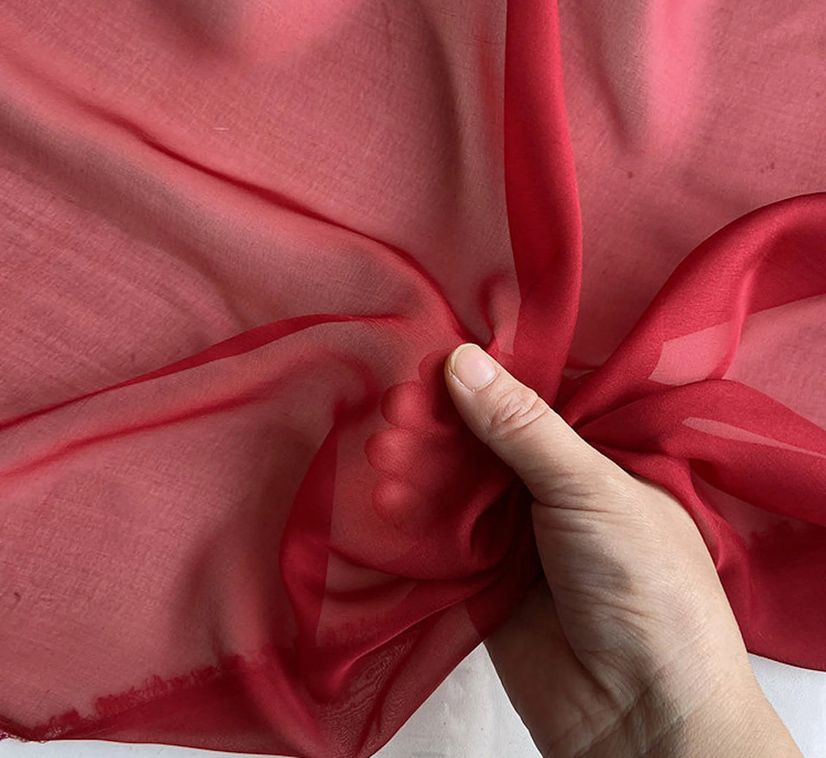 A hand is folding a piece of sheer, reddish-pink silk chiffon fabric.