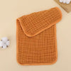 A stack of three orange quilted muslin baby burp cloths resting on a light beige surface.