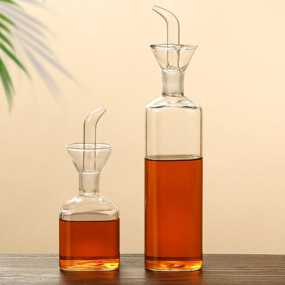 A clear borosilicate glass oil and vinegar dispenser filled with amber liquid, sitting on a wooden surface.