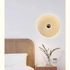 A modern, round, translucent yellow LED wall sconce with a touch dimming feature mounted on a white wall above a wooden bed.