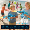 A young boy holds and fires a blue electric water gun at the beach with a smiling woman nearby.