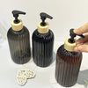 A set of two black glass refillable soap dispensers with wooden pump tops and a natural fiber cork stopper.