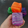 Une main tient un cube de jeu en gel anti-stress rayé violet et rose, à côté de deux autres cubes colorés.