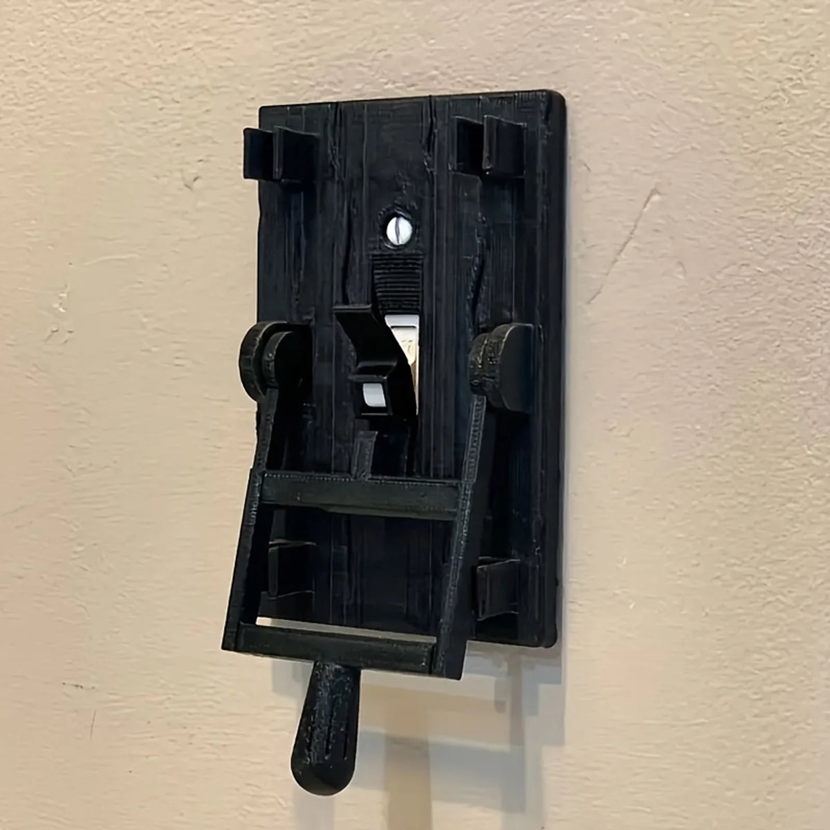 A black decorative light switch cover with a flip handle mounted on a beige wall.
