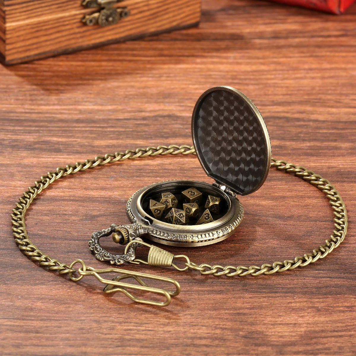A gold-toned chain watch case with an open lid revealing seven metal polyhedral dice on a wooden surface.