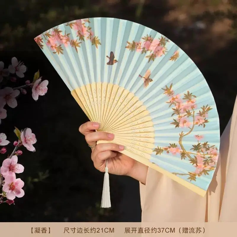 A hand holds an open bamboo and silk folding fan with a light blue background and pink floral design.