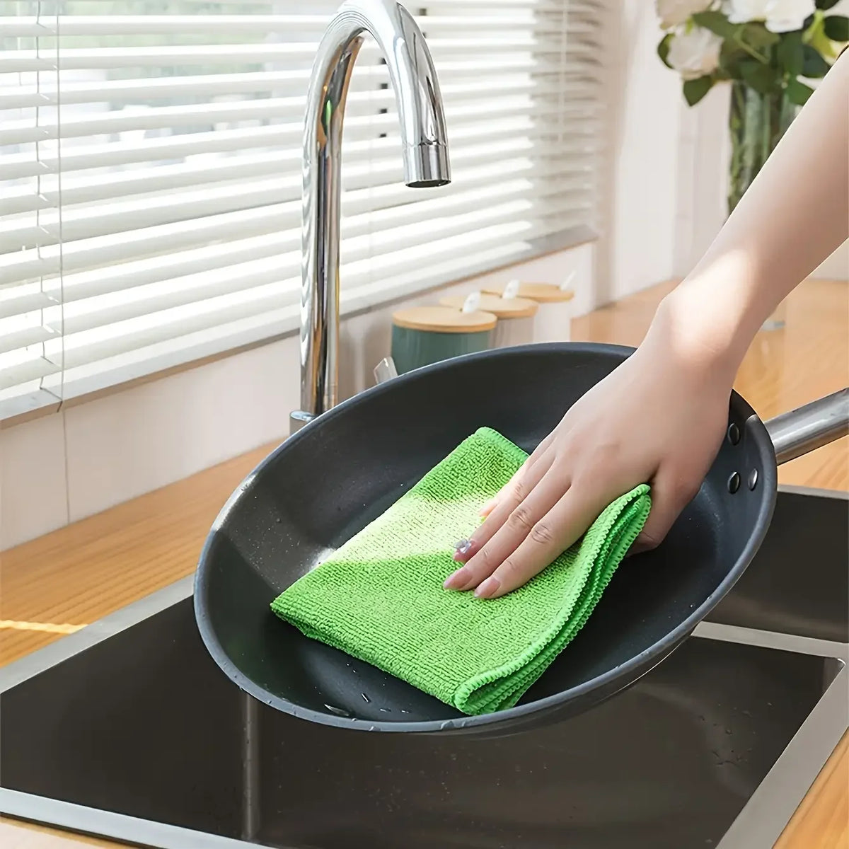A hand is using a bright green microfiber cleaning cloth to wipe a black frying pan on a stovetop.