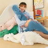 A woman is sitting on a bed surrounded by various soft plush toys including blue and pink dragon-shaped pillows.