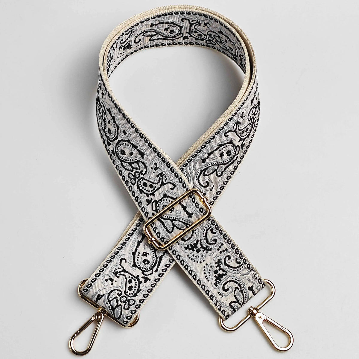 A beige and black paisley jacquard strap for a bag, featuring an antique gold buckle.