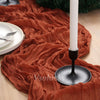 A terracotta crinkle gauze table runner draped over a wooden surface with a white candle and dark stand.