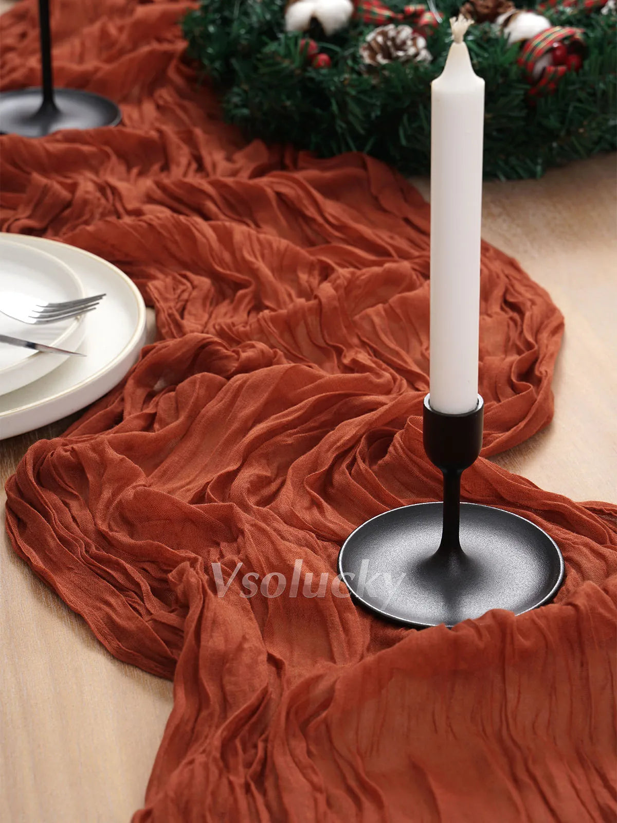 A terracotta crinkle gauze table runner draped over a wooden surface with a white candle and dark stand.