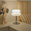 A modern cloud-shaped bedside table lamp with a white shade sits on a light wood surface next to a potted plant.