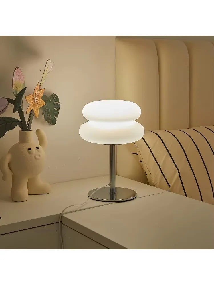 A modern cloud-shaped bedside table lamp with a white shade sits on a light wood surface next to a potted plant.