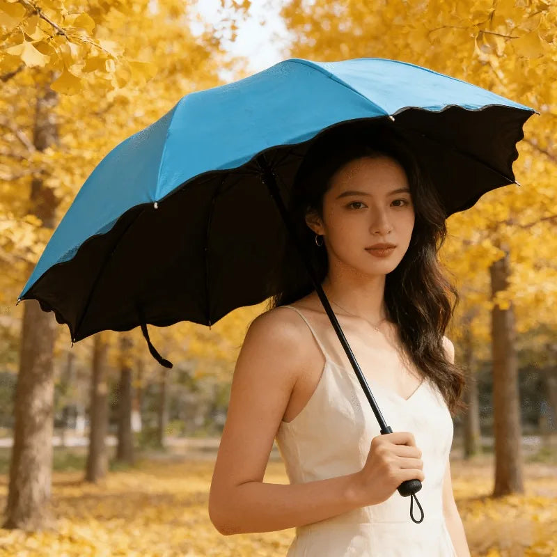 A woman holds a black umbrella under a bright blue umbrella in an outdoor park with yellow autumn foliage.