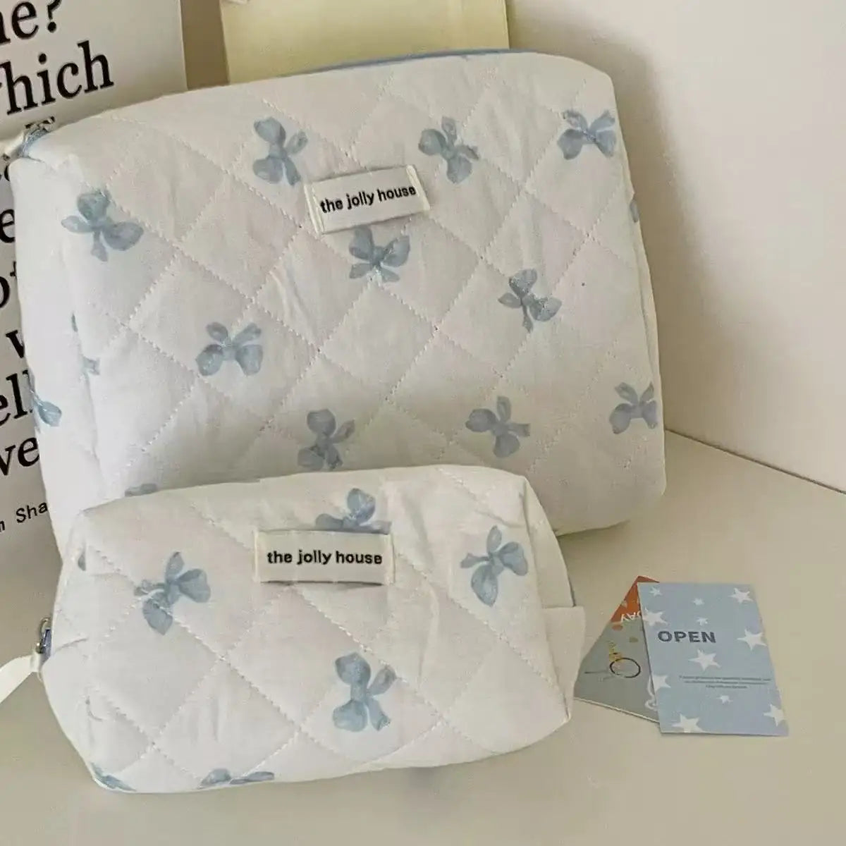 A white quilted cosmetic bag with blue bow print and the jolly house logo, shown in a studio setting.