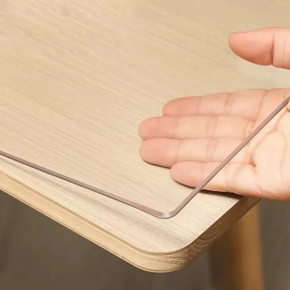 A hand is holding a metal table protector, made of clear PVC, over a wooden table surface.