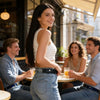 A young woman wearing a white tank top and jeans smiles while sitting at an outdoor cafe table.