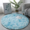 A bright blue shaggy circle area rug placed on a light gray floor with a potted plant and a sofa in the background.