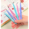 A hand holds several colorful macaron-shaped craft tweezers used for scrapbooking and journaling.