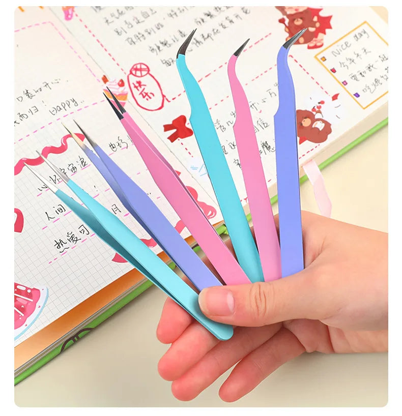 A hand holds several colorful macaron-shaped craft tweezers used for scrapbooking and journaling.