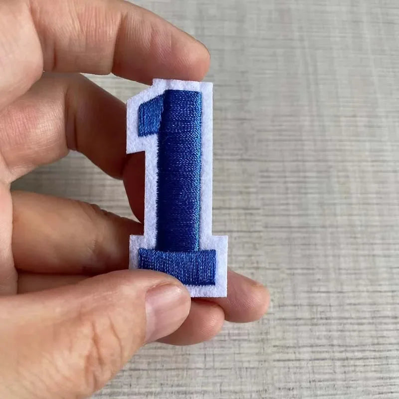 A hand holds a blue iron-on 3D number patch shaped like the numeral one.
