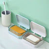 A white transparent soap dish with a drainage tray and lid is shown next to a toothbrush on a light green wall.