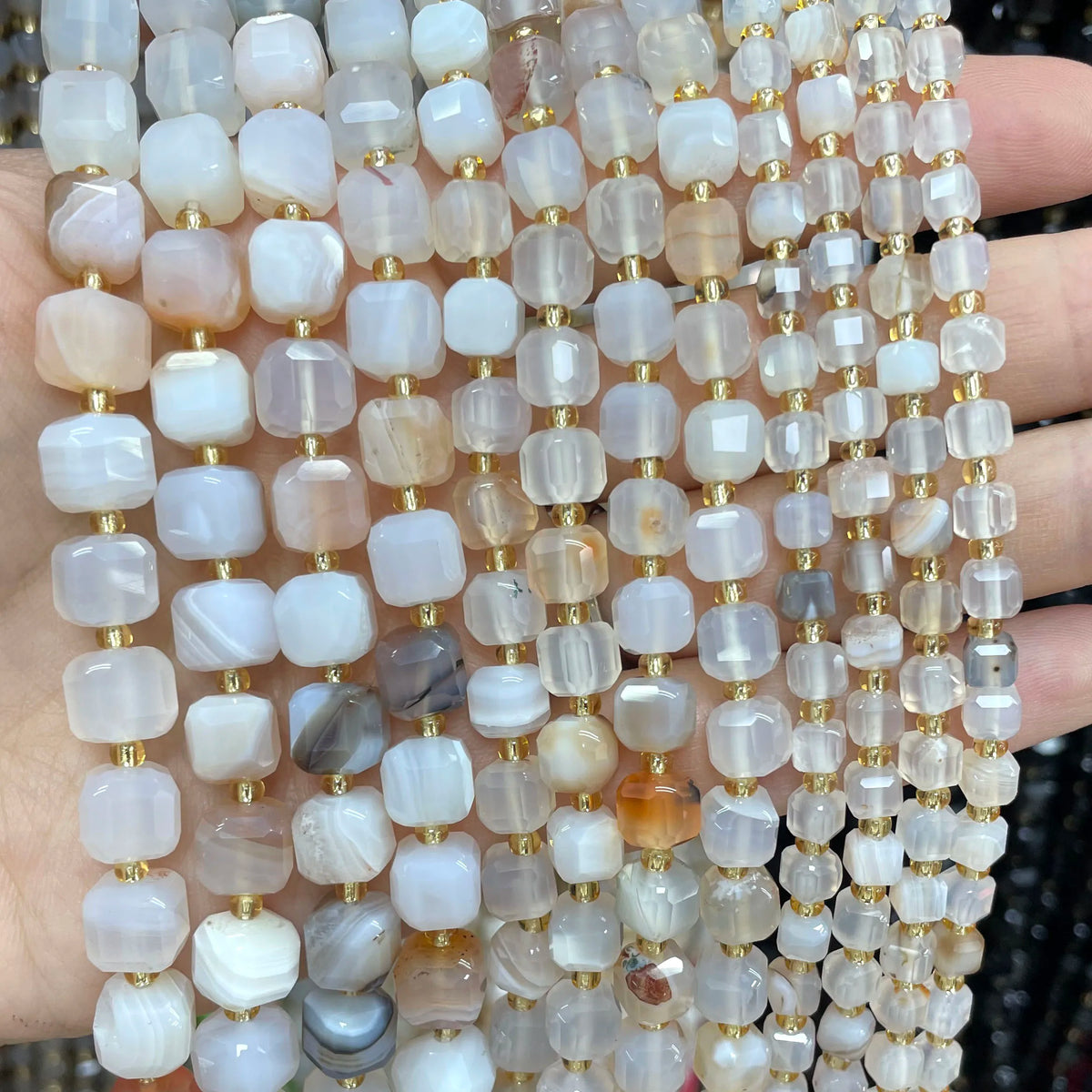 A hand holds a long strand of square faceted natural stone beads in various shades of pink, beige, and clear quartz.