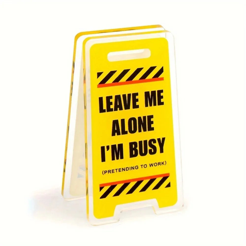 A yellow acrylic desk sign with black diagonal stripes and the text LEAVE ME ALONE I'M BUSY PRETENDING TO WORK