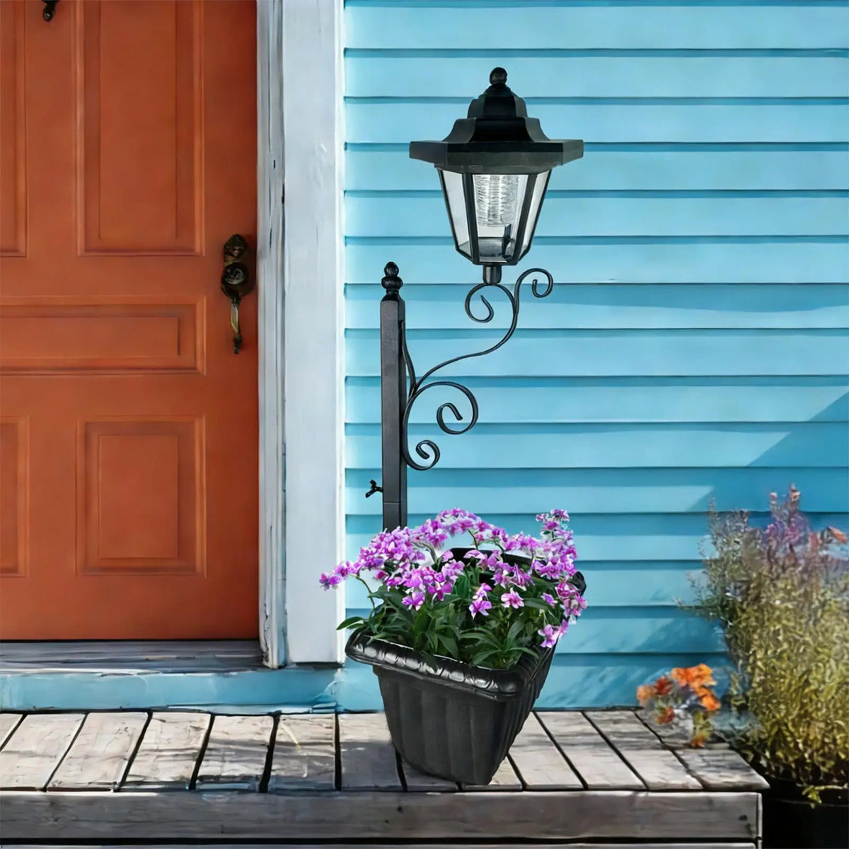 A black wrought iron solar-powered planter light with purple flowers sits on a wooden deck next to an orange door.
