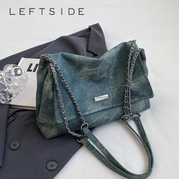 Dunkeltealiges Lederhandtasche mit silberner Kettchenriemen neben einem dunkelgrauen Blazer.