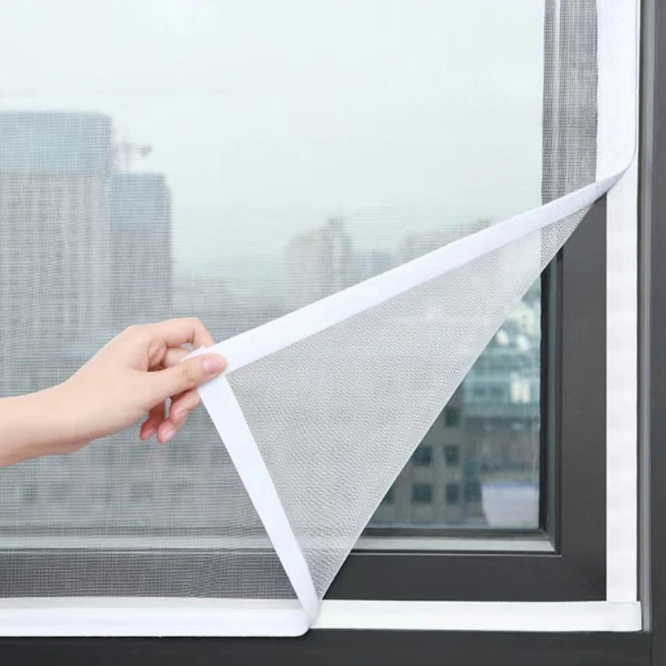 A hand is holding a white self-adhesive window screen mesh with a zipper, demonstrating the product for use on a window.
