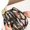 A hand is holding a clear PVC toiletry bag with black trim, showing various toiletries inside.
