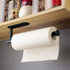 A white roll of toilet paper is displayed on a black adhesive stainless steel rack mounted under a wooden shelf.
