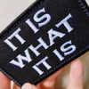 Black embroidered tactical morale patch with white text reading It is what it is being held by a hand