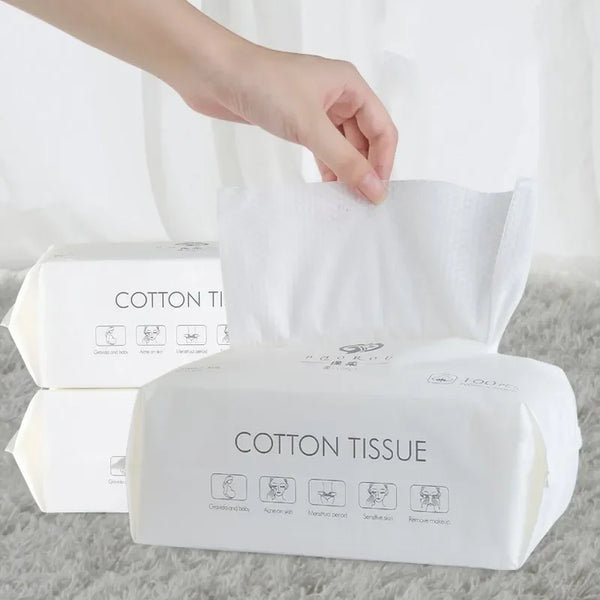 A hand is opening a white package of soft disposable cotton facial tissues on a gray surface.