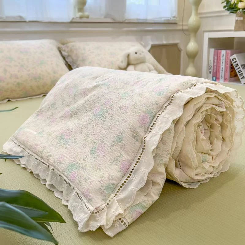 A rolled floral washed cotton gauze summer quilt with delicate lace edging resting on a light green surface.