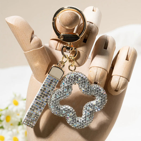 A close-up of a rhinestone clover keychain is held between fingers against a light background.