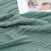 A soft sage green muslin throw blanket draped over a surface next to a white card.