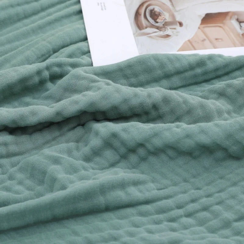 A soft sage green muslin throw blanket draped over a surface next to a white card.