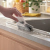 A hand is using a white multi-purpose crevice cleaning brush to clean a window track on a countertop.