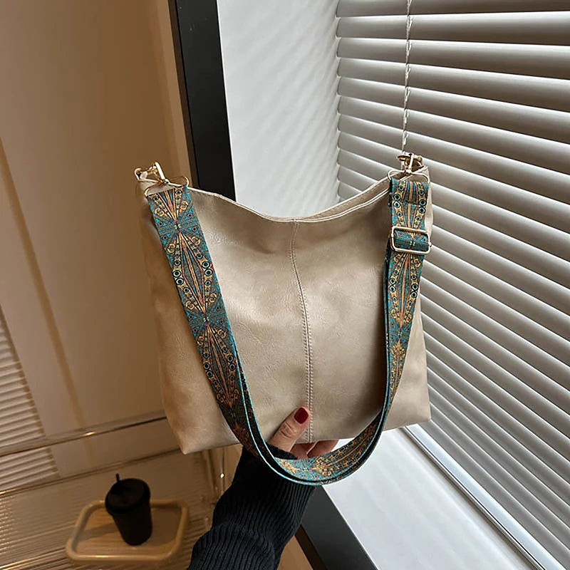 Sac à l'épaule beige avec une bretelle géométrique large aux motifs bleu sarcelle et or, accroché devant un store de fenêtre.