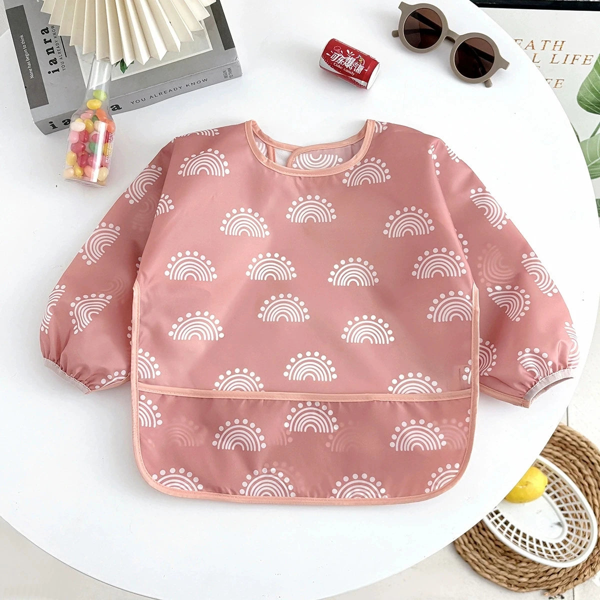 A pink waterproof long-sleeve baby feeding smock with a white rainbow pattern displayed on a white surface.