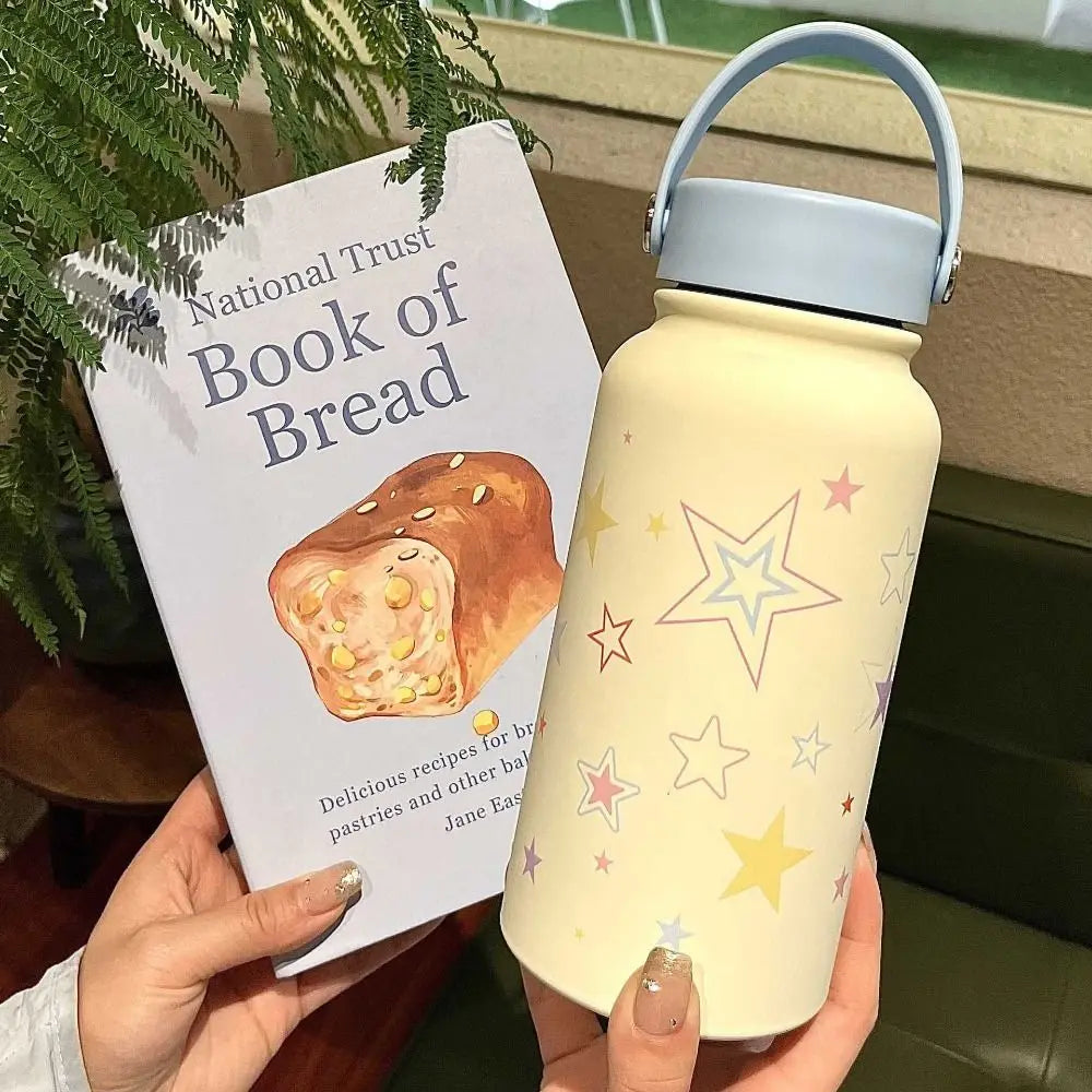 A hand holds a light yellow insulated tumbler with a blue lid and star pattern, next to a book titled National Trust Book of 