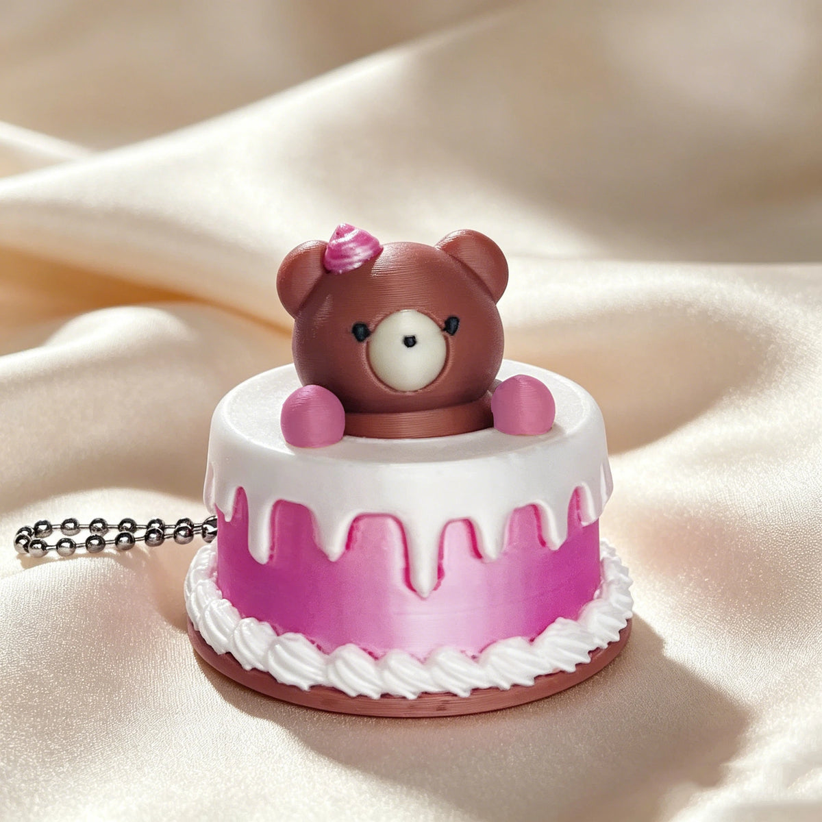 A round pink and brown cartoon bear cake with white frosting and pink icing details, resting on a satin fabric.