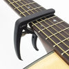 A black adjustable guitar capo with a built-in bridge pin puller is shown on a wooden guitar neck.