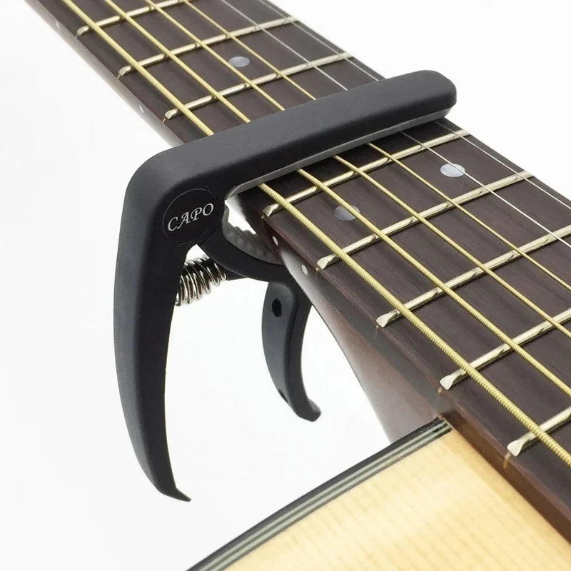 A black adjustable guitar capo with a built-in bridge pin puller is shown on a wooden guitar neck.
